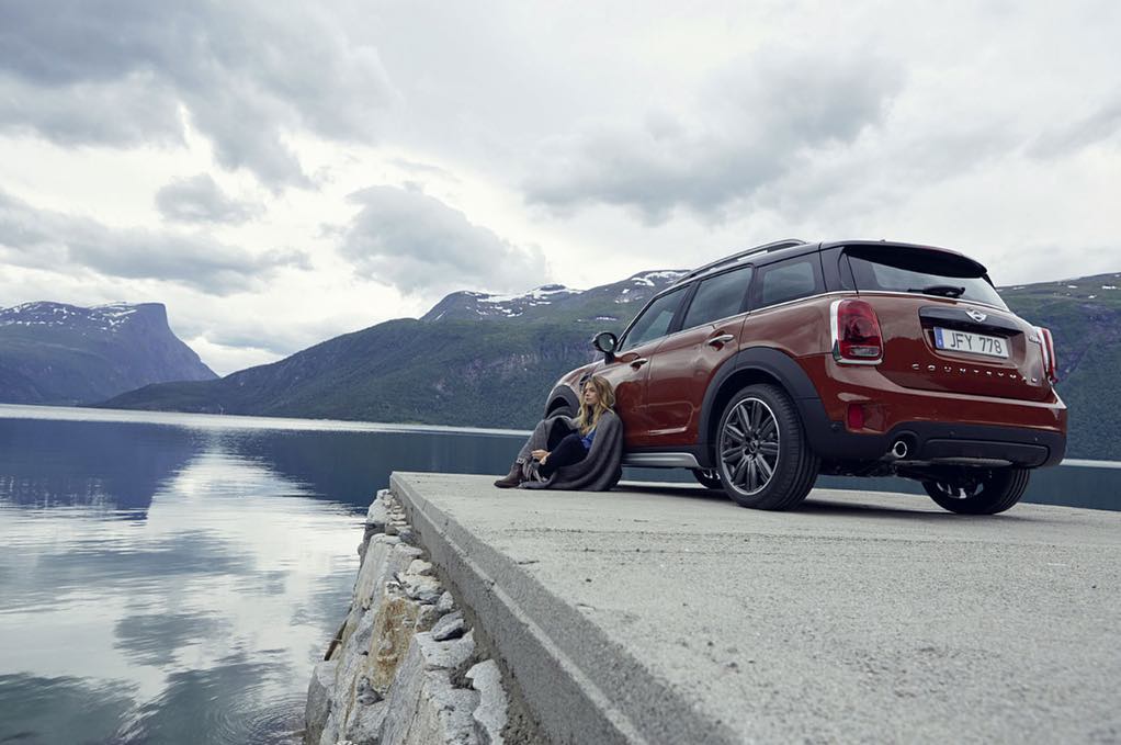 An auburn Mini Countryman SUV parked by a lake in Norway, with a young woman sitting on a rock wall beside the car, mountains visible in background.