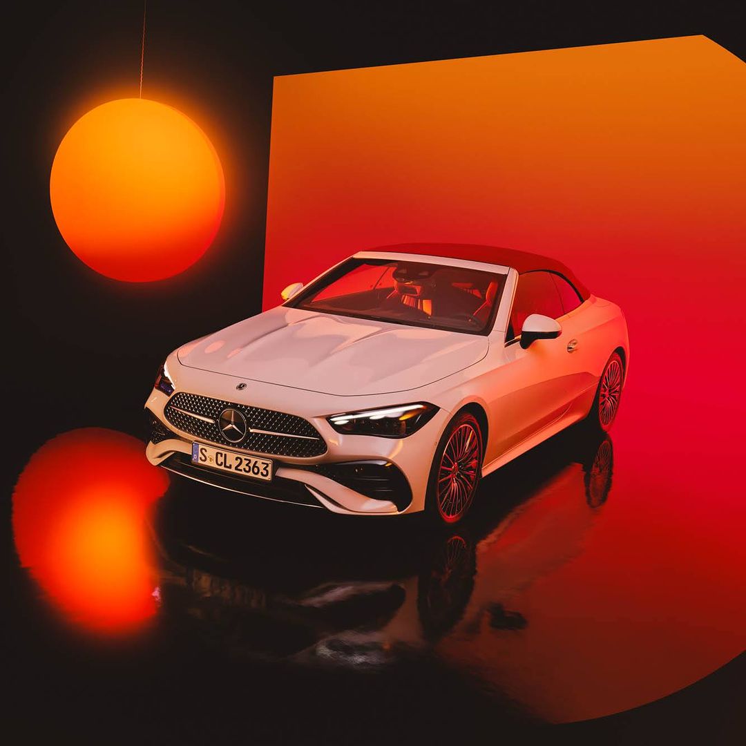 White Mercedes-Benz convertible car on display in a studio with orange lighting and black reflective floor.