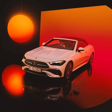 White Mercedes-Benz convertible car on display in a studio with orange lighting and black reflective floor.