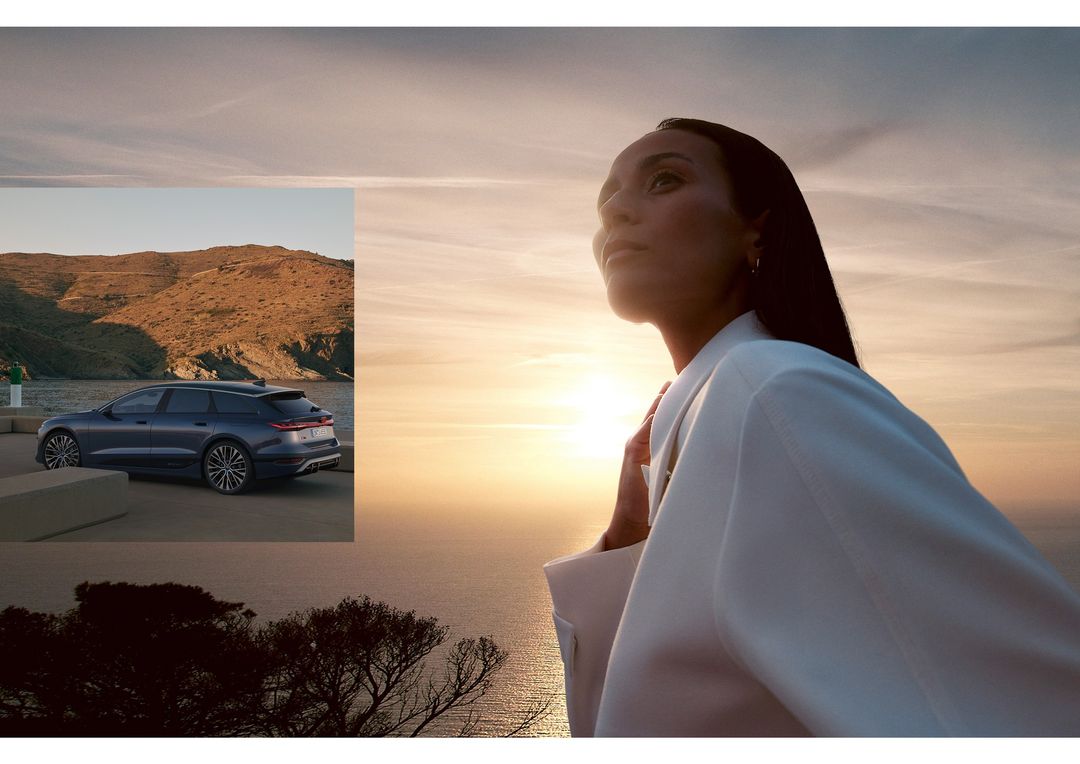 Woman in a white blazer looks out over the ocean at sunset alongside a gray Audi station wagon on a coastal road.