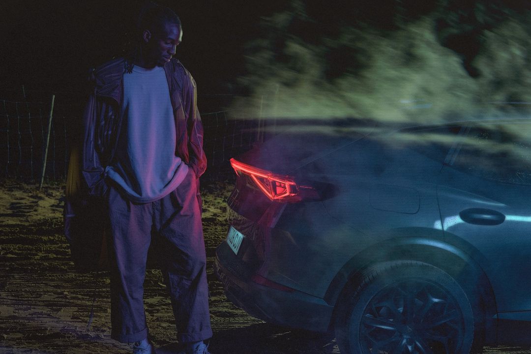 Night automotive fashion shoot featuring a Black male adult in casual wear standing near a dark grey coupe, set against a dark outdoor backdrop.