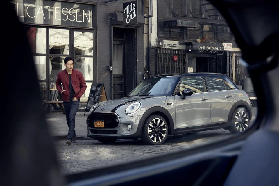Silver Mini Cooper parked on a city street with a man walking nearby. Street signs and building facades form the urban background.