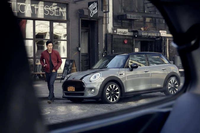 Silver Mini Cooper parked on a city street with a man walking nearby. Street signs and building facades form the urban background.