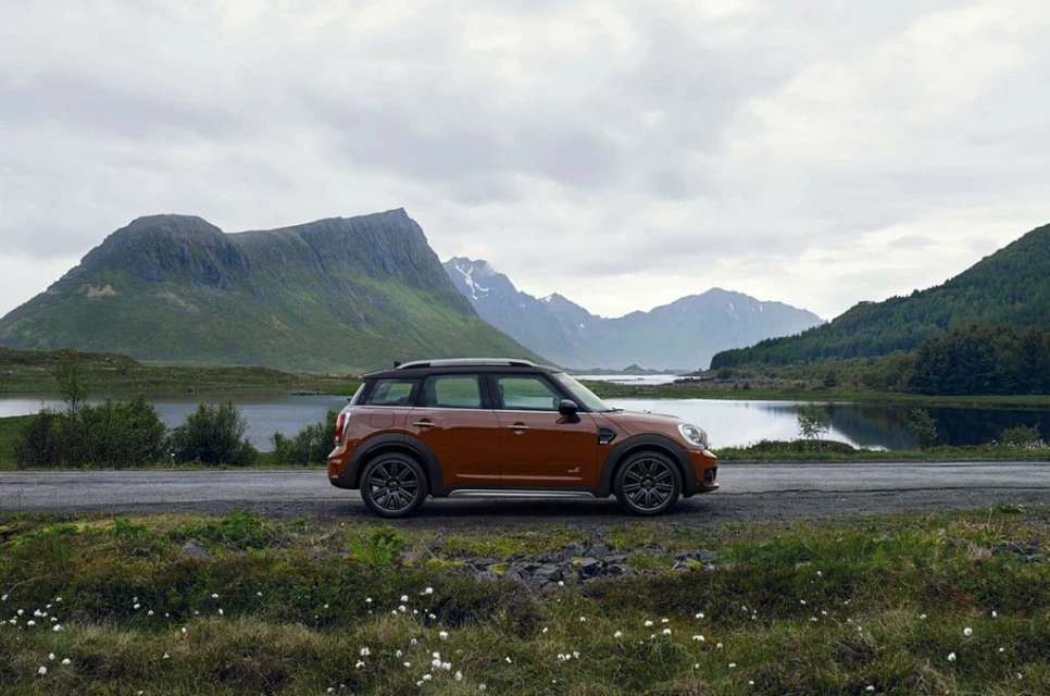 Orange Mini Countryman parked on the roadside with a backdrop of mountains, a lake, and a cloudy sky.
