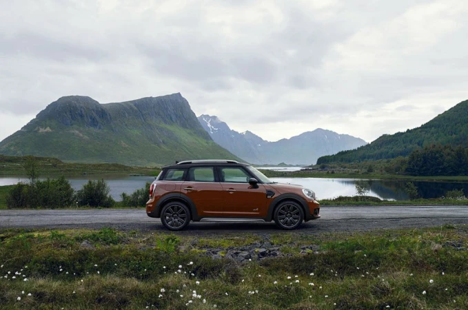 Orange Mini Countryman parked on the roadside with a backdrop of mountains, a lake, and a cloudy sky.