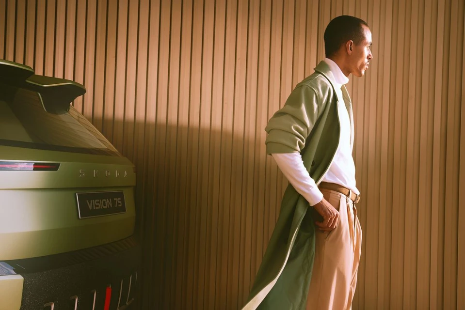 A man in a light green coat stands near the rear of a green Skoda Vision 7S concept car against a light brown, vertically slatted wall.