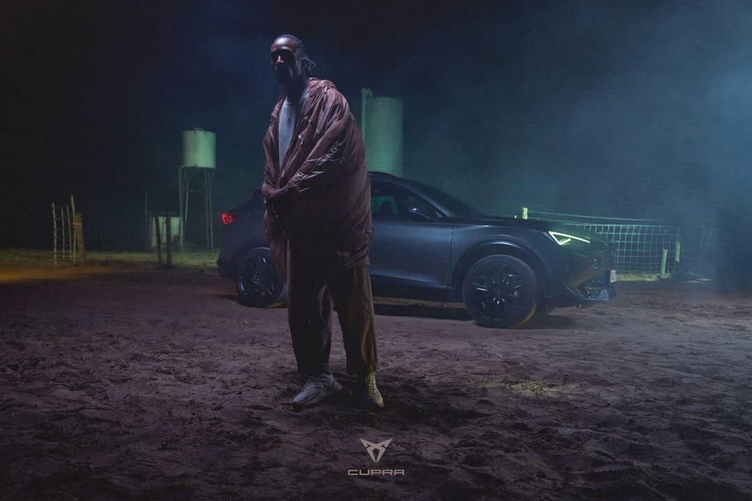 Man in orange jacket poses near dark grey Cupra car in a nighttime outdoor setting with dirt ground and water tower backdrop.