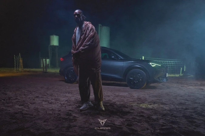 Man in orange jacket poses near dark grey Cupra car in a nighttime outdoor setting with dirt ground and water tower backdrop.