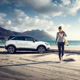 Woman walking on a sandy beach carrying a yoga mat next to a parked white crossover SUV with mountains in the background