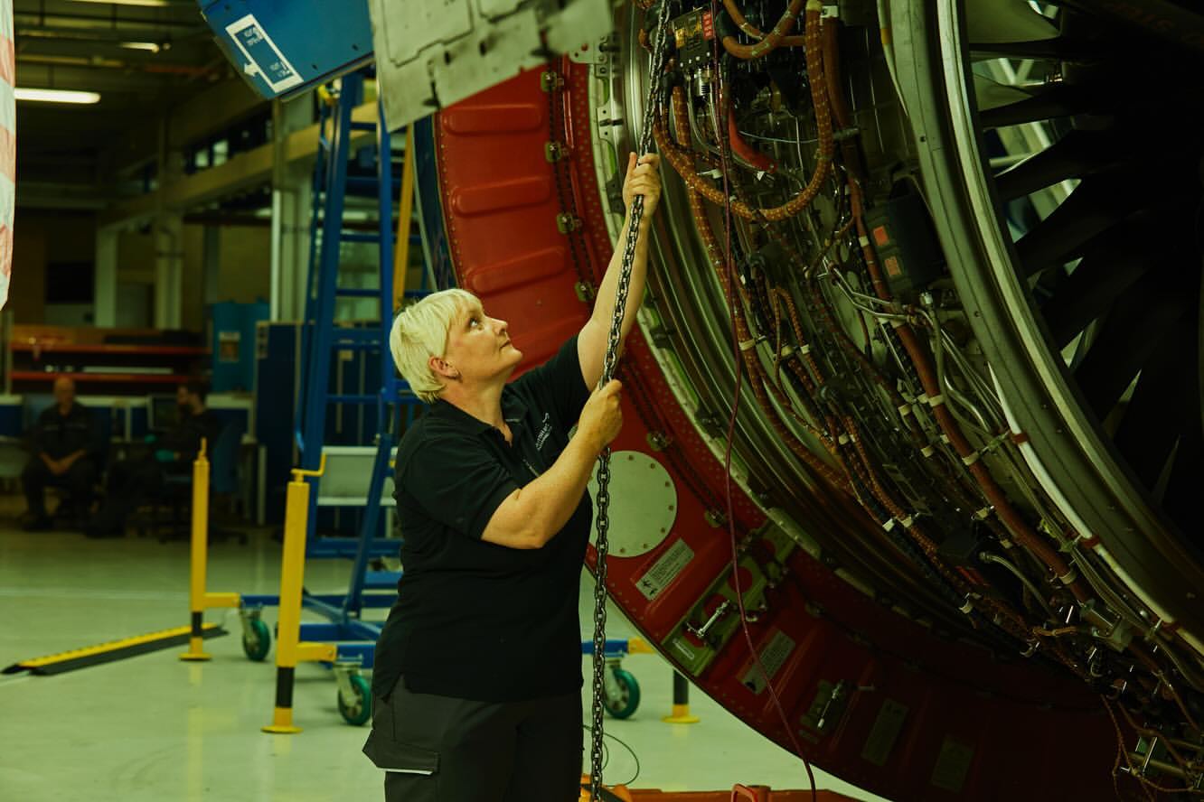 Adult woman working on an aircraft engine in an industrial setting, using a chain; showcasing industrial maintenance in muted lighting.