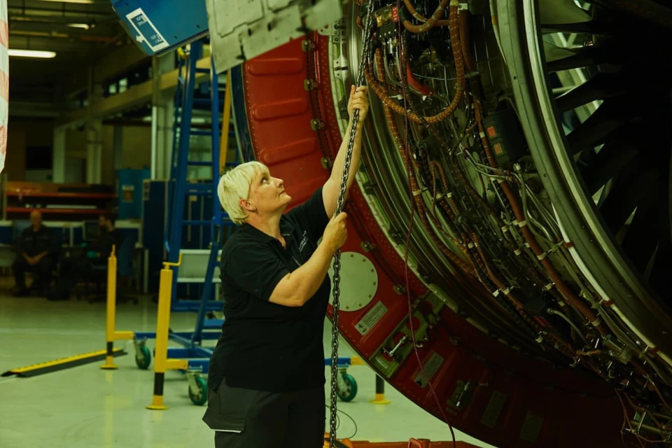 Adult woman working on an aircraft engine in an industrial setting, using a chain; showcasing industrial maintenance in muted lighting.