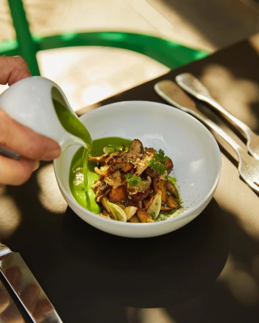 Overhead view of green soup poured over mushrooms and potatoes in a white bowl, served with silverware on a black surface.