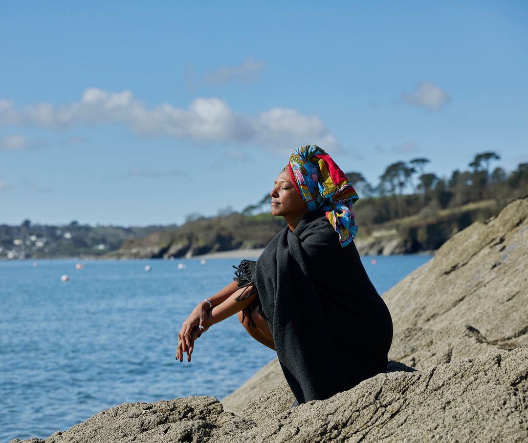 Woman sitting on rocks by the sea with a colorful headscarf, wrapped in a dark shawl enjoying the sun on a clear, sunny day.