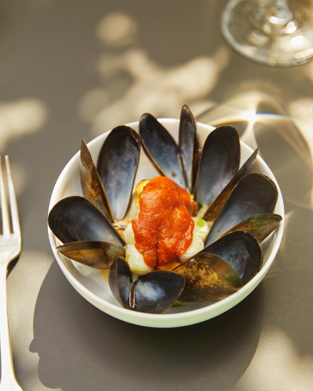 Overhead shot of a white bowl with mussels in their shells and tomato sauce on a grey surface, next to a metal fork.