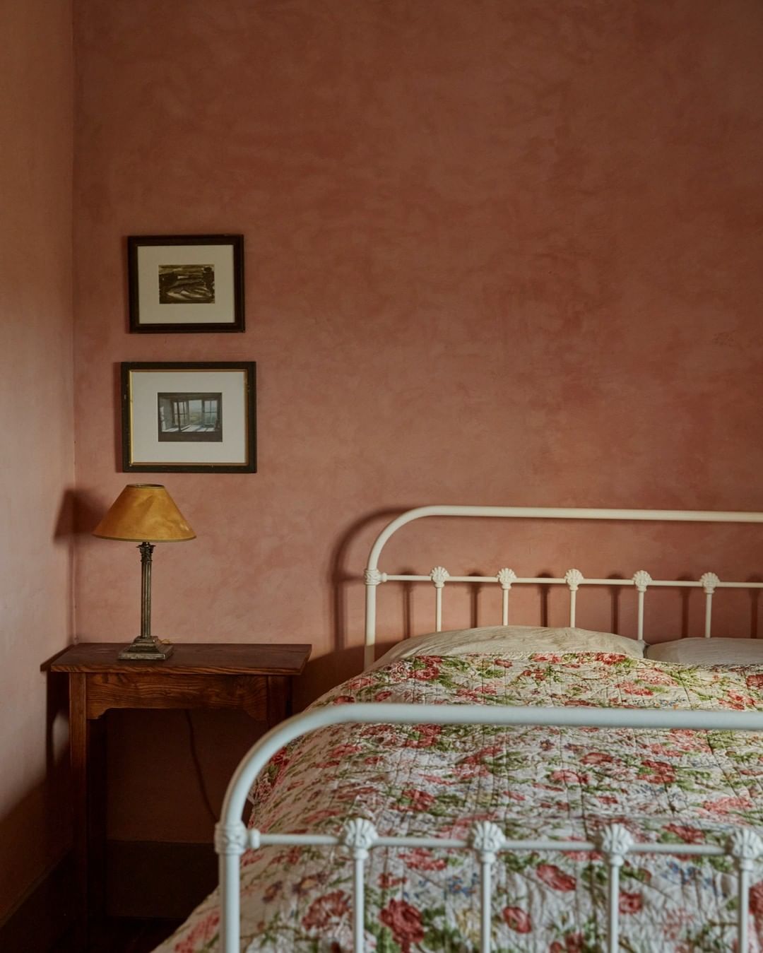 Bedroom interior featuring a white iron bed with floral quilt, a wooden bedside table with a lamp, and artwork on a textured pink wall.
