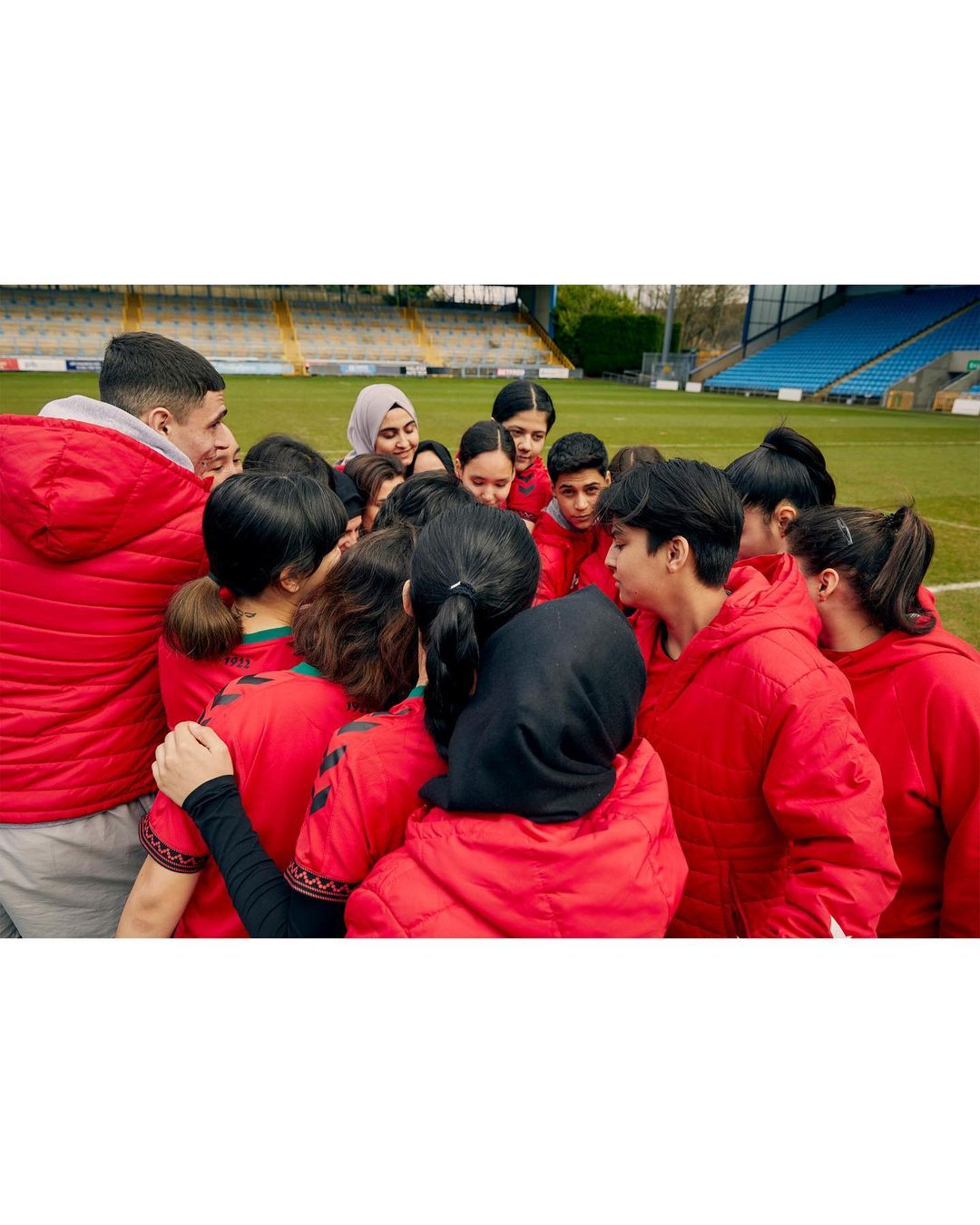A group of young adults and teens huddled together on a soccer field, wearing matching red and green sportswear, with stadium seating visible.