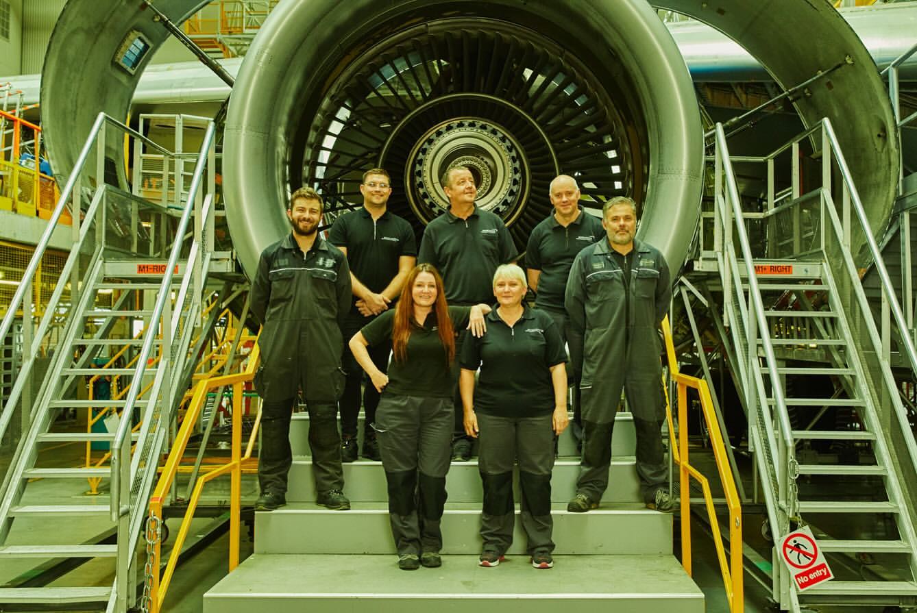 Group of aircraft engineers standing in front of a jet engine inside of an airplane hangar, wearing dark clothing, and posing for a picture.