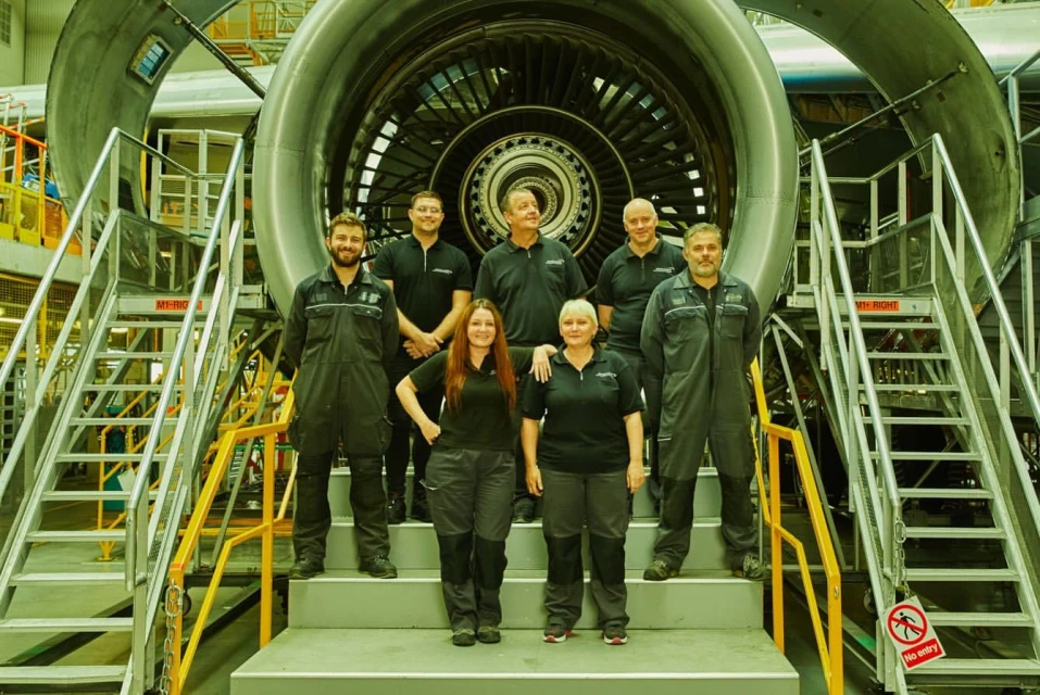 Group of aircraft engineers standing in front of a jet engine inside of an airplane hangar, wearing dark clothing, and posing for a picture.