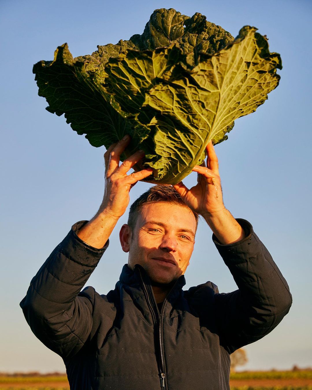 Adult male holding a large, leafy vegetable over his head outside in a rural field under a blue sky during daylight hours.