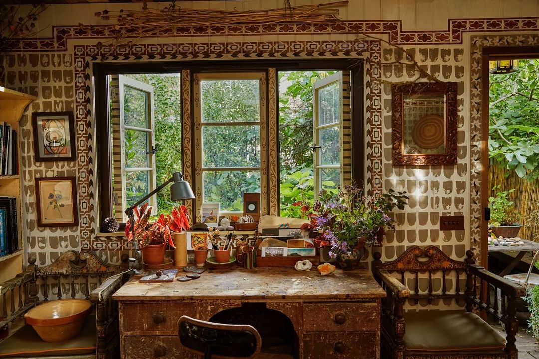 Interior of an artist's studio with a rustic wooden desk, antique chair, potted plants, and natural light streaming through a multi-paned window.