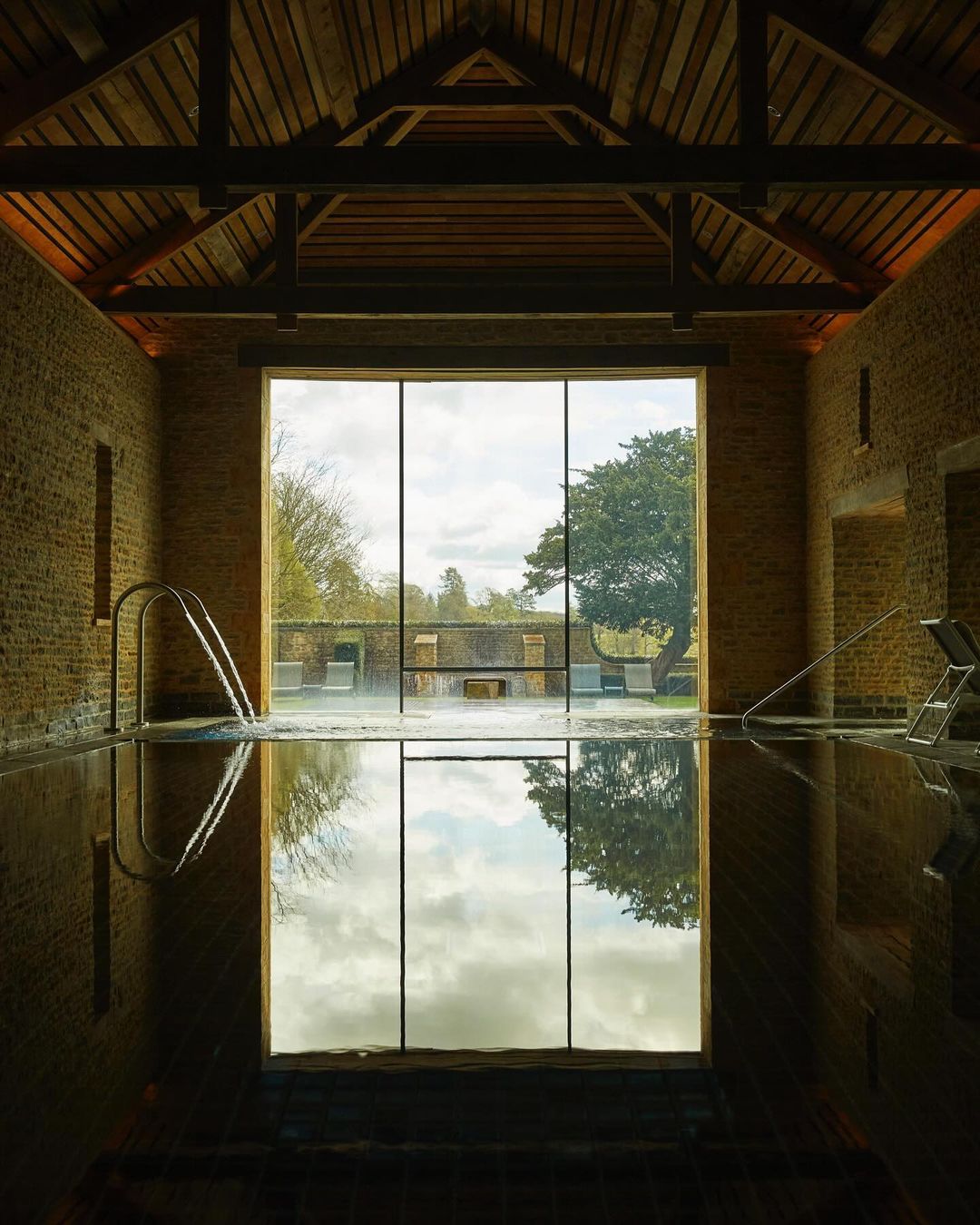 Indoor swimming pool with stone walls, dark water reflecting the sky, and a window showing trees and landscape on the opposite wall.