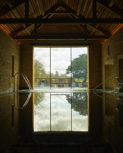 Indoor swimming pool with stone walls, dark water reflecting the sky, and a window showing trees and landscape on the opposite wall.