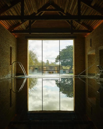 Indoor swimming pool with stone walls, dark water reflecting the sky, and a window showing trees and landscape on the opposite wall.