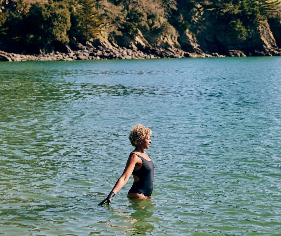 Adult woman in a black swimsuit stands in teal-green water, arm extended. A tree-covered shoreline is visible in the background.