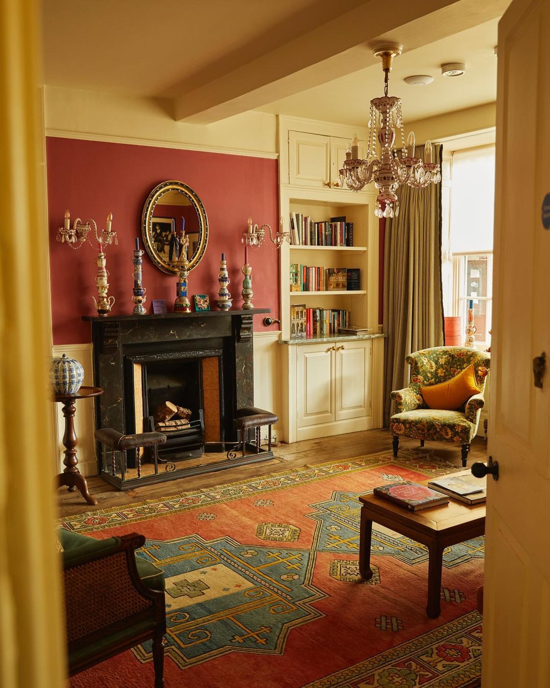 Warm interior view of a living room featuring a fireplace, patterned rug, chandelier, and a doorway leading to another room.