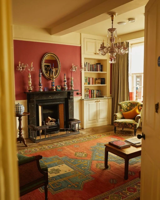 Warm interior view of a living room featuring a fireplace, patterned rug, chandelier, and a doorway leading to another room.