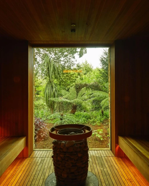 Indoor view of a sauna room with wood plank walls and floors, with a heater, and a garden view through a window.