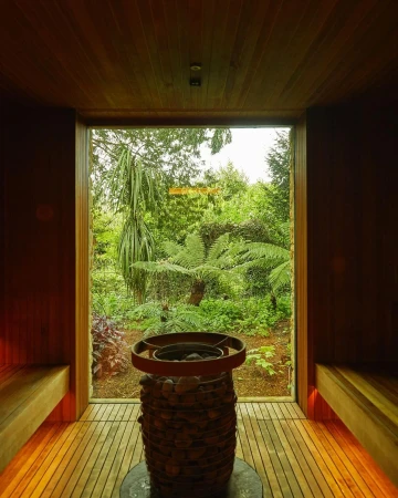 Indoor view of a sauna room with wood plank walls and floors, with a heater, and a garden view through a window.