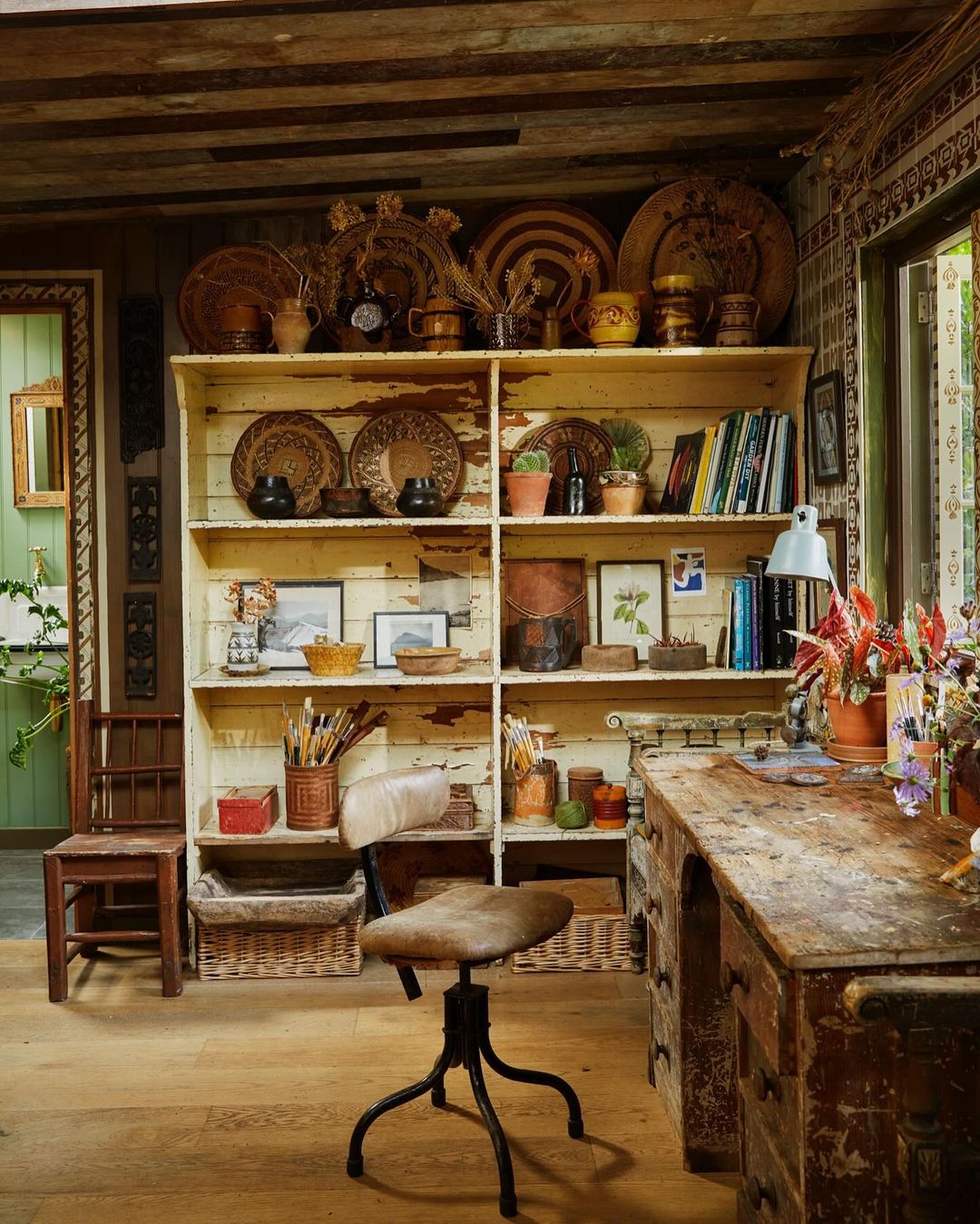 Rustic interior with a wooden desk, antique chair, and shelves filled with decorative items and books in a warm, inviting space.