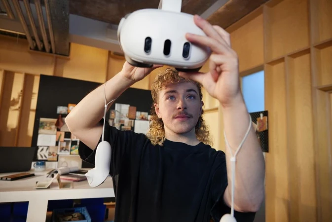 Young adult male with curly hair is indoors, holding a white VR headset in his hands, standing in a brightly lit home studio setting.