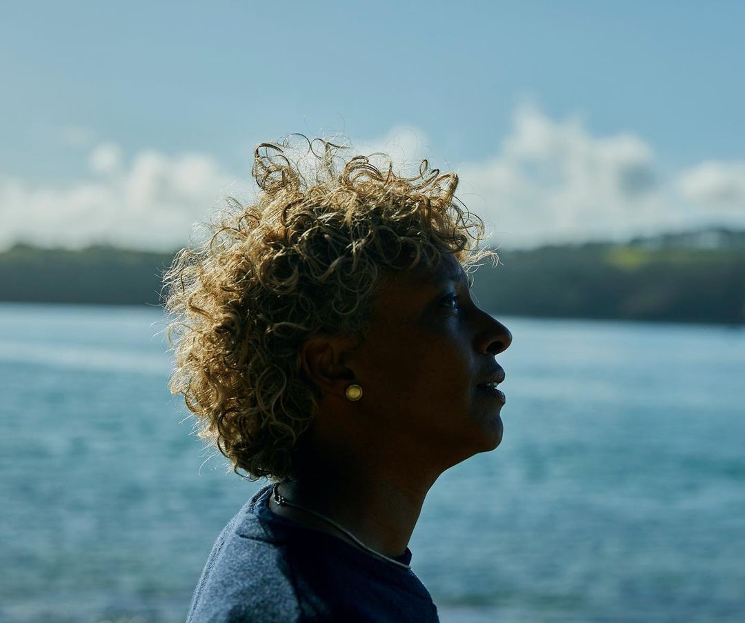 Profile of an adult woman with curly hair looking up outdoors against a background of the ocean, sky, and vegetation on the shoreline.
