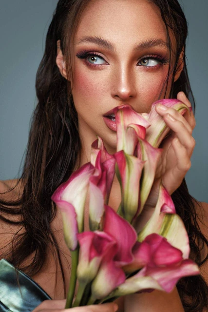 Close-up of a woman with wet hair holding pink calla lilies, makeup, and blue background in a beauty and fashion headshot.
