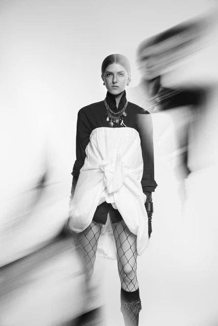 Monochrome portrait of a woman in a black top, white skirt, and fishnet stockings, against a blurred white background. Fashion.