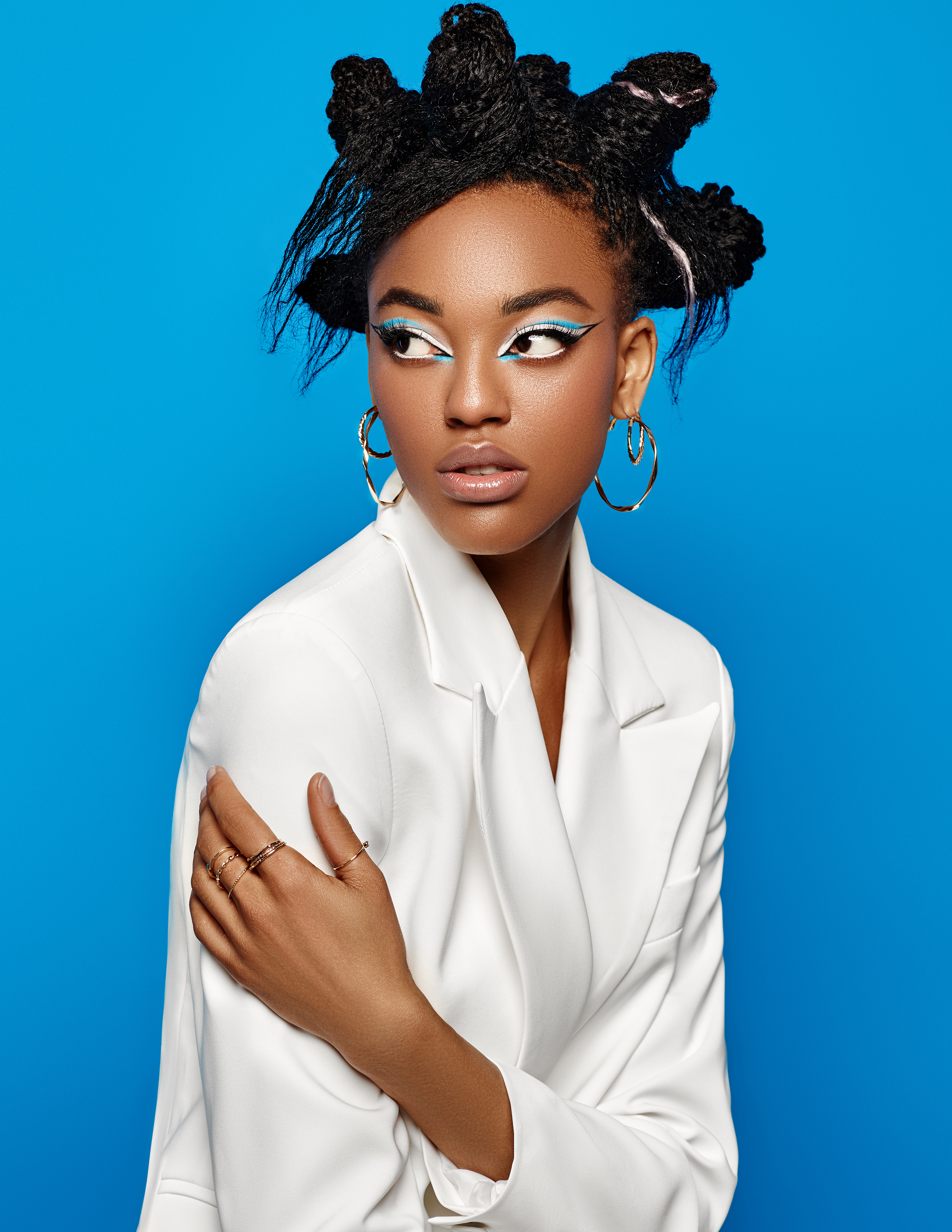 Young woman with unique hairstyle and bright makeup poses in a white blazer against a blue background, showcasing a modern fashion look.