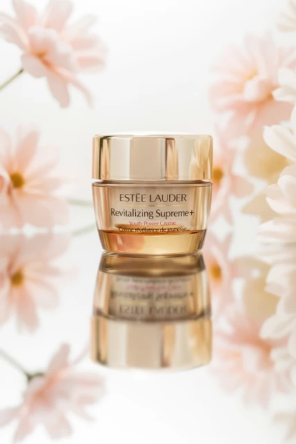 Estee Lauder Revitalizing Supreme+ cream jar on a reflective surface surrounded by light pink daisy flowers, set against a white background.