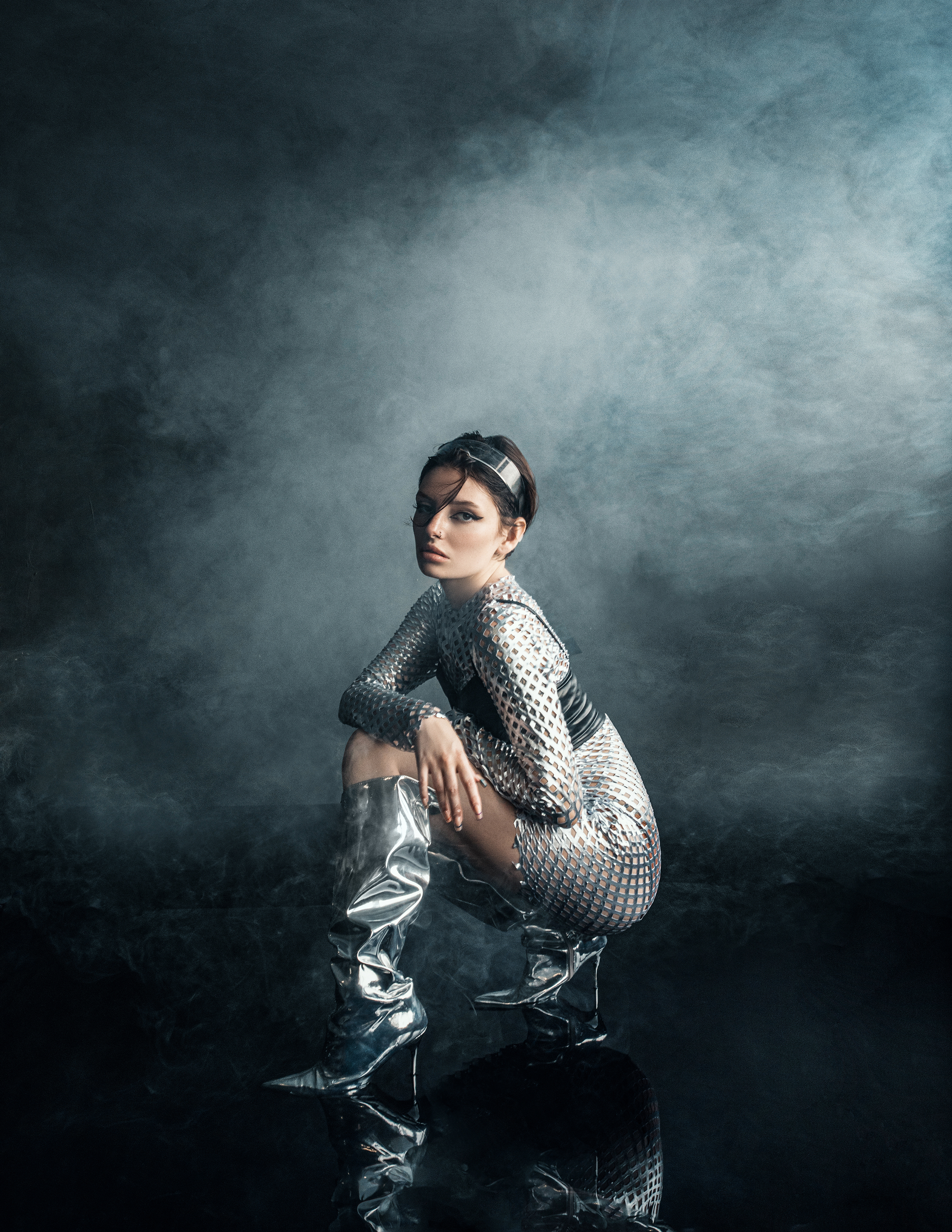 Woman in shiny, futuristic outfit and boots poses in studio with smoke. The pose is squatting on reflective surface. Studio shot.