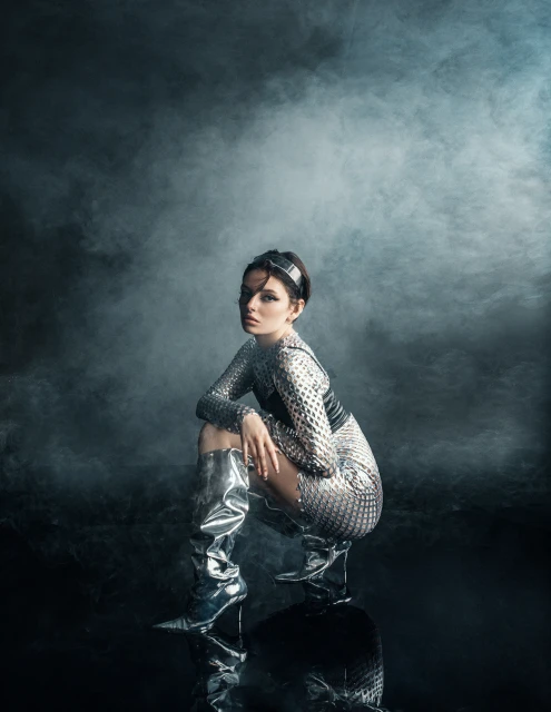 Woman in shiny, futuristic outfit and boots poses in studio with smoke. The pose is squatting on reflective surface. Studio shot.