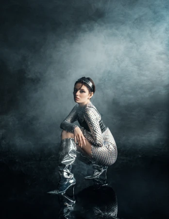 Woman in shiny, futuristic outfit and boots poses in studio with smoke. The pose is squatting on reflective surface. Studio shot.