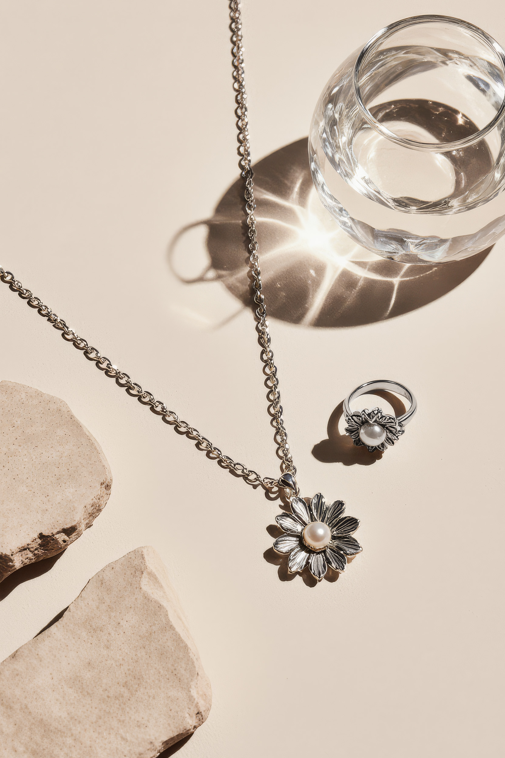 Silver necklace with flower pendant, matching ring with pearl, and glass of water on a beige surface, casting soft shadows.