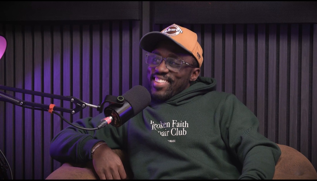 A man with glasses and a hat smiles next to a microphone while wearing an green hoodie in front of a dark vertical slat wall.