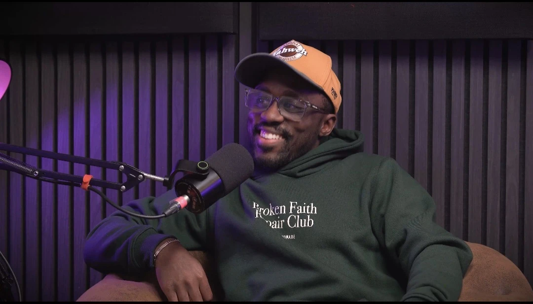 A man with glasses and a hat smiles next to a microphone while wearing an green hoodie in front of a dark vertical slat wall.