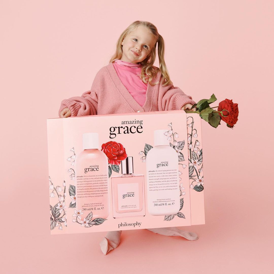 Young girl holding a pink Philosophy gift box with beauty products and a red rose against a pink background in a studio setting.