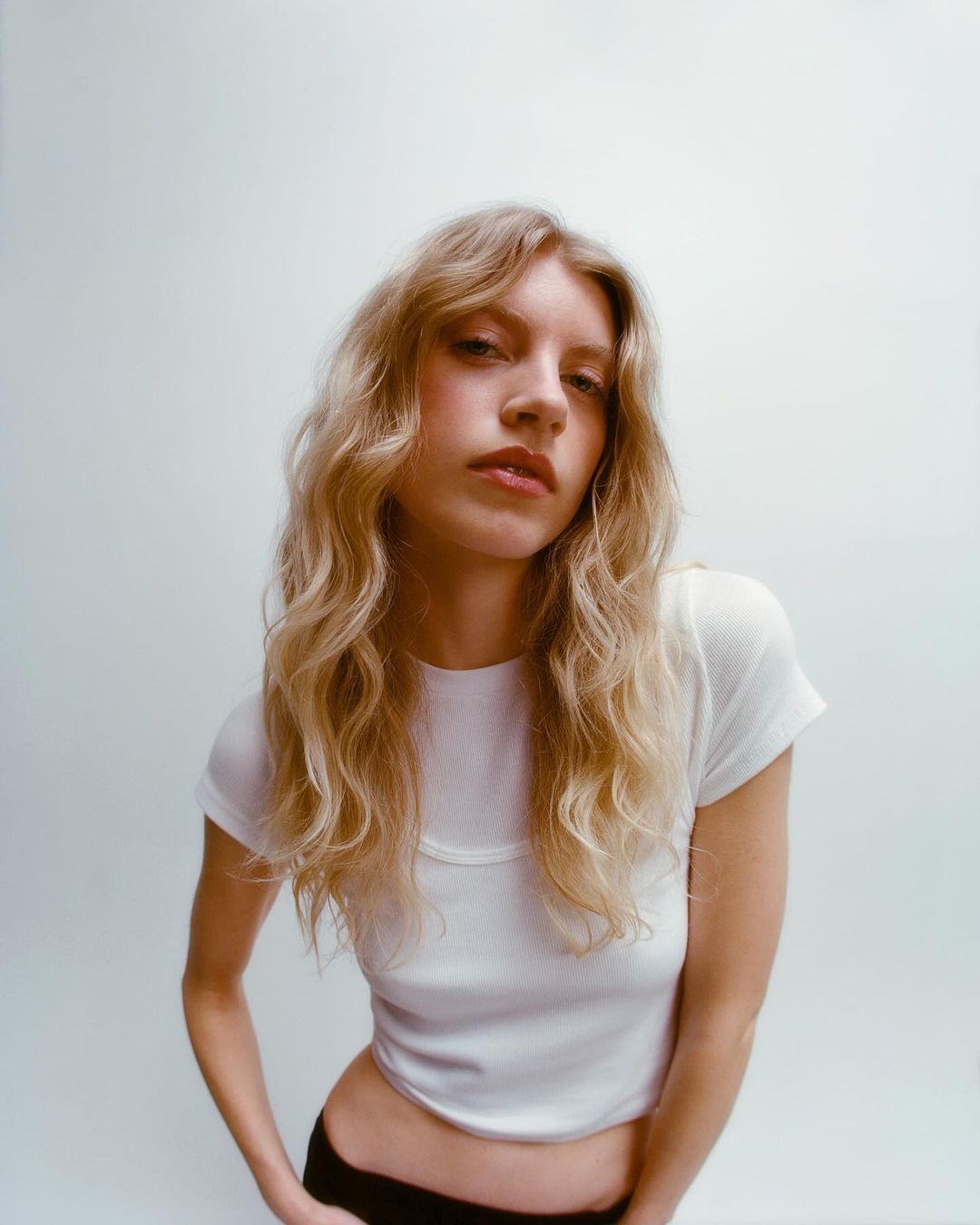 Blonde woman with wavy hair poses in a white short-sleeve shirt against a bright background in a studio portrait. Fashion shot.