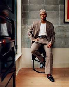 Adult male in tan suit and matching head covering sits on stool in front of brick wall, posing indoors near window.