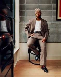 Adult male in tan suit and matching head covering sits on stool in front of brick wall, posing indoors near window.
