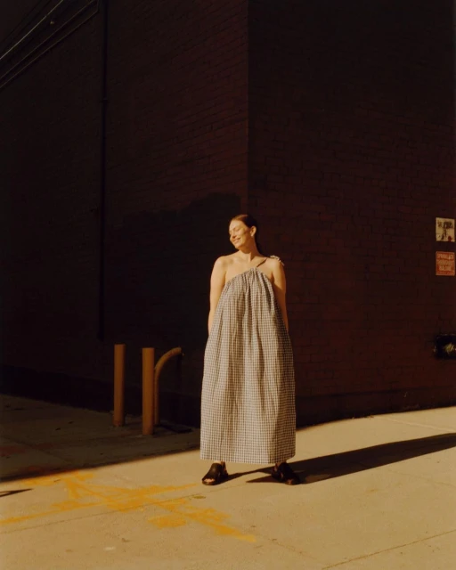 Woman wearing a gingham dress stands in sunlight against a dark wall, looking off to the side in an outdoor setting, fashion.